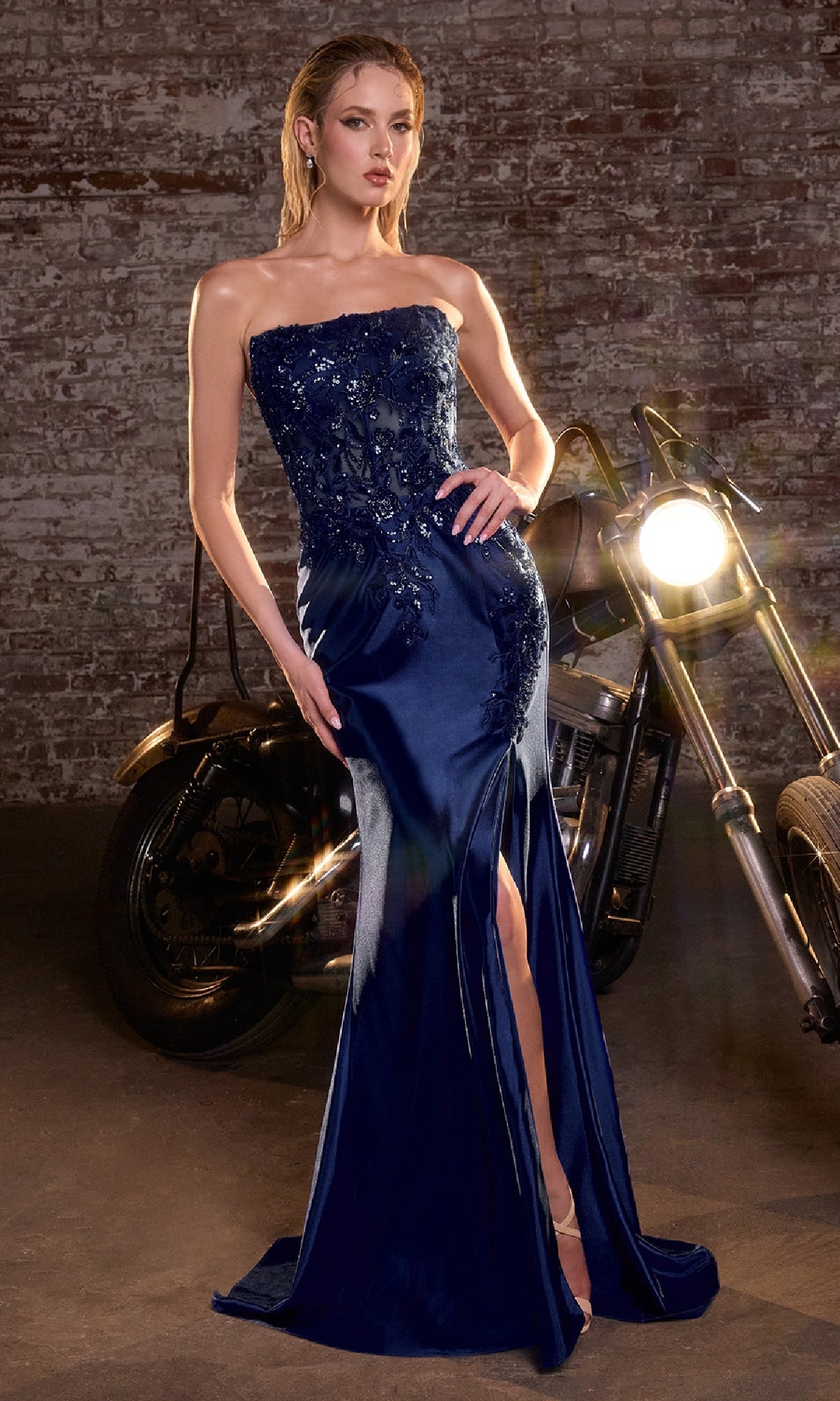 Woman in a strapless navy blue evening gown with a high slit, standing next to a motorcycle against a brick wall.